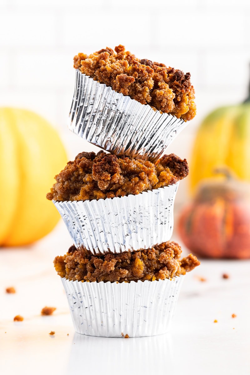 Three Keto Pumpkin Cream Cheese Muffins stacked on top of each other in silver muffin liners, displayed on a white counter. The muffins are topped with a crumbly streusel, and decorative pumpkins are softly blurred in the background, enhancing the autumn vibe.