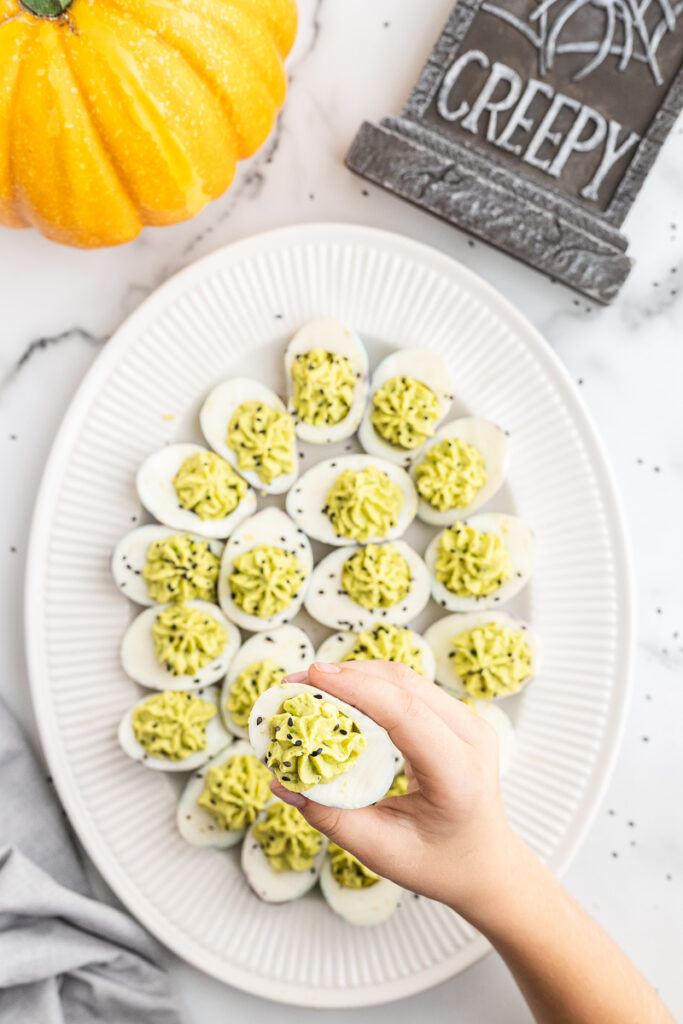 Overhead photo of Halloween Deviled Eggs on a white platter with a child's hand holding one deviled egg up for the camera.