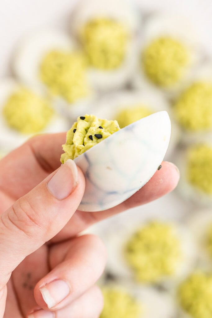 Someone holding a Halloween Deviled Eggs above the platter of them on a white marble counter.