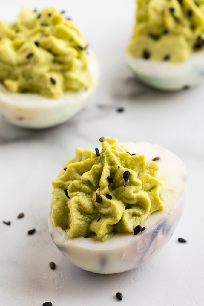 Closeup photo of three Halloween Deviled Eggs on a white marble counter with black sesame seeds sprinkled on and around them.