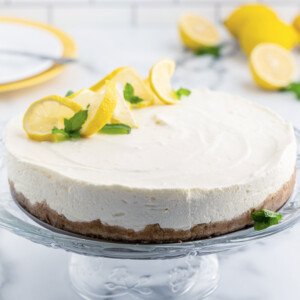 Keto no-bake lemon cheesecake on a glass cake plate with lemons in the background on a white marble counter.