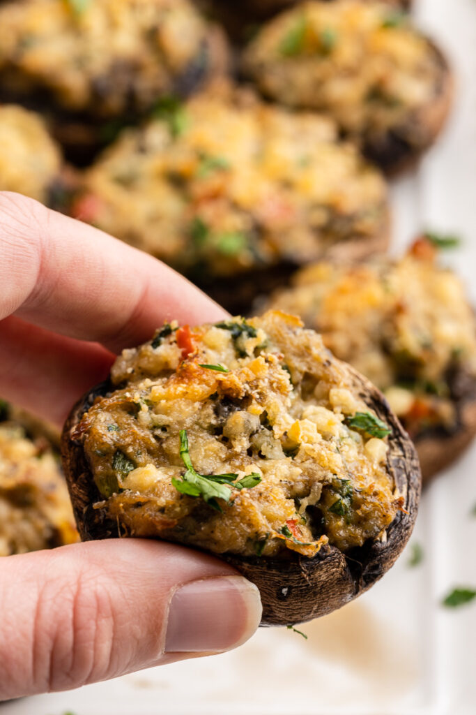 Close up of a hand holding a Keto Crab Stuffed Mushroom above a bronze baking sheet full of them.