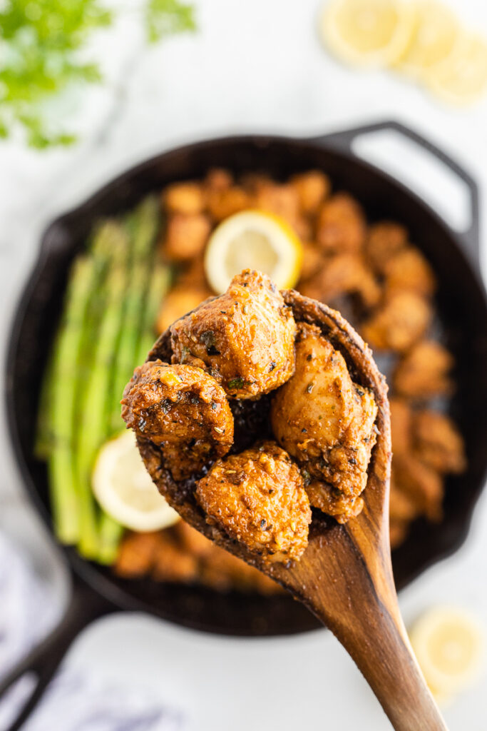 Close-up photo of a wooden serving spoon full of Lemon Garlic Chicken Bites with the remaining chicken and asparagus in the cast iron skillet below.