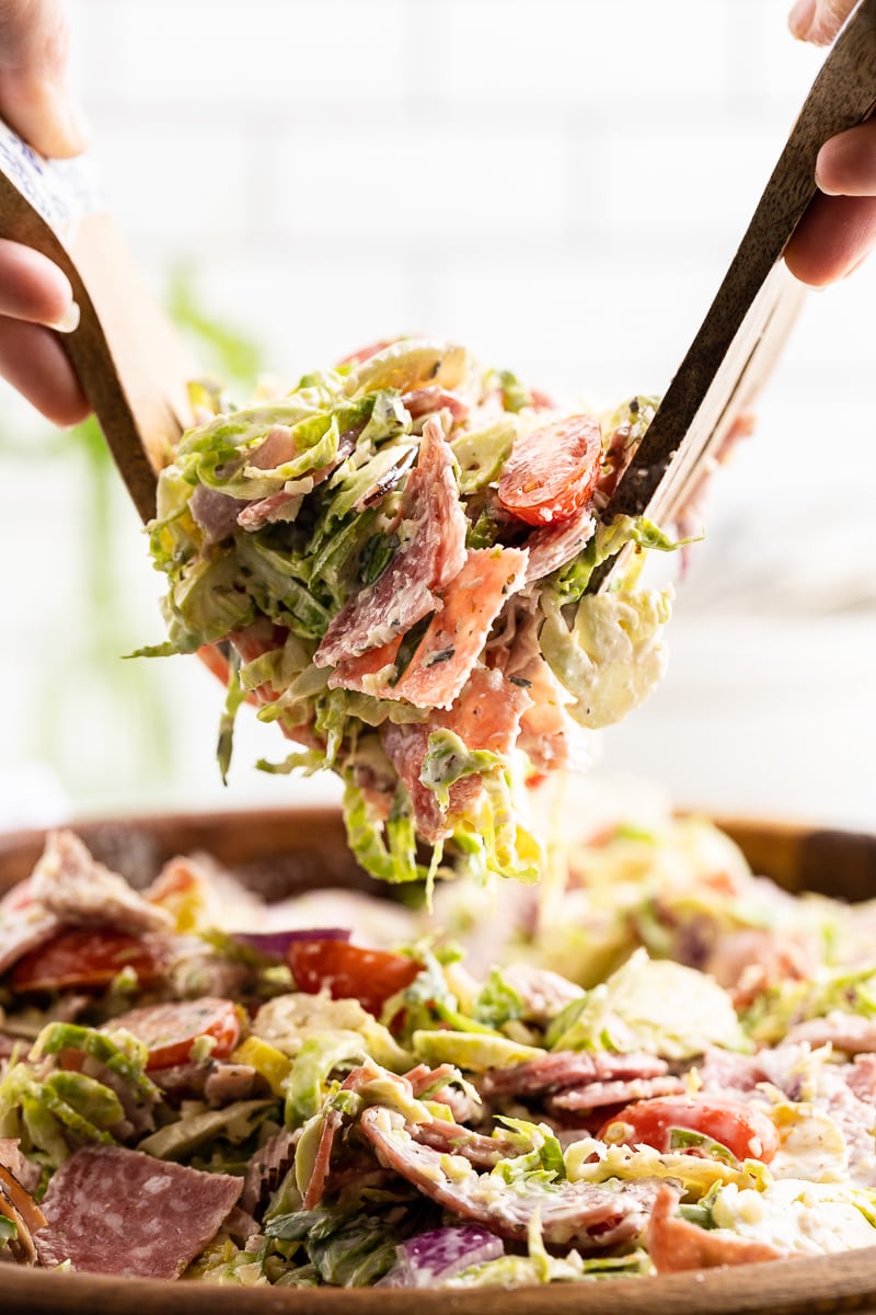 Close up of Italian Grinder Salad in a wooden salad bowl, served with wooden salad tongs, featuring shredded Brussels sprouts, salami, ham, provolone cheese, red onion, and cherry tomatoes, dressed with homemade dressing.