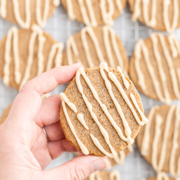 A hand holding a Keto Chai Sugar Cookie close to the camera with a metal cooking rack full of them in the background.