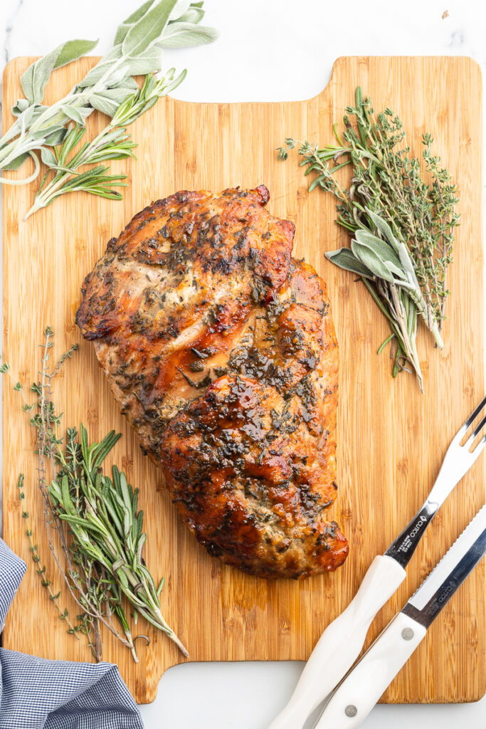 Maple Mustard Roasted Turkey Breast on a wooden cutting board with fresh herbs around it.