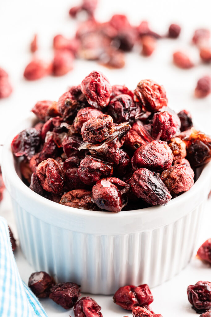 Homemade Sugar-Free Dried Cranberries in a white ramekin with some scattered on the white countertop beside it.