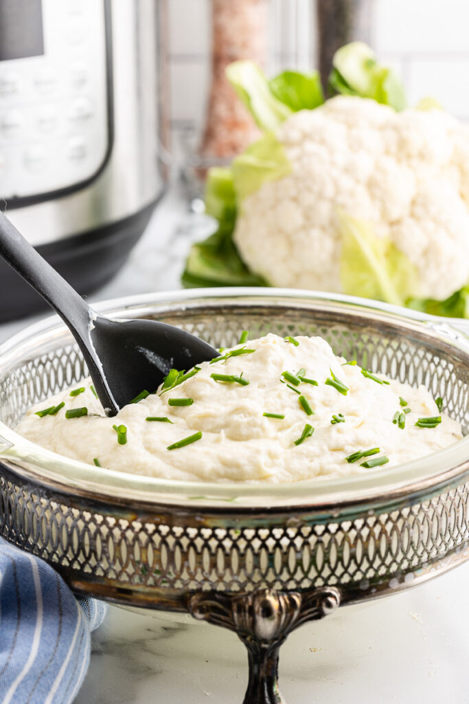 Instant Pot Creamy Garlic Mashed Cauliflower in a glass serving dish with a head of cauliflower and the Instant Pot in the background.