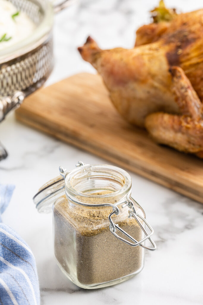 A glass spice jar full of Homemade Poultry Seasoning on a white marble counter with a cooked whole chicken on a wooden cutting board in th ebackground.