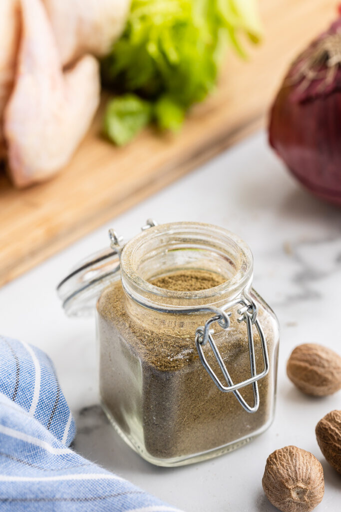 A glass spice jar full of Homemade Poultry Seasoning on a white marble counter with whole nutmeg, a red onion, and a whole chicken in the background.