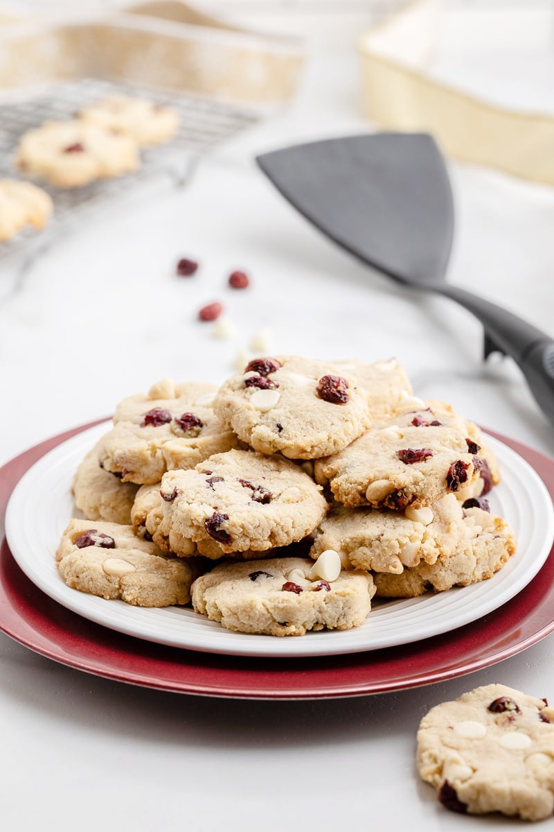 Freshly baked keto white chocolate cranberry cookies on a white plate with a cooling rack full of cookies and a black spatula in the background.