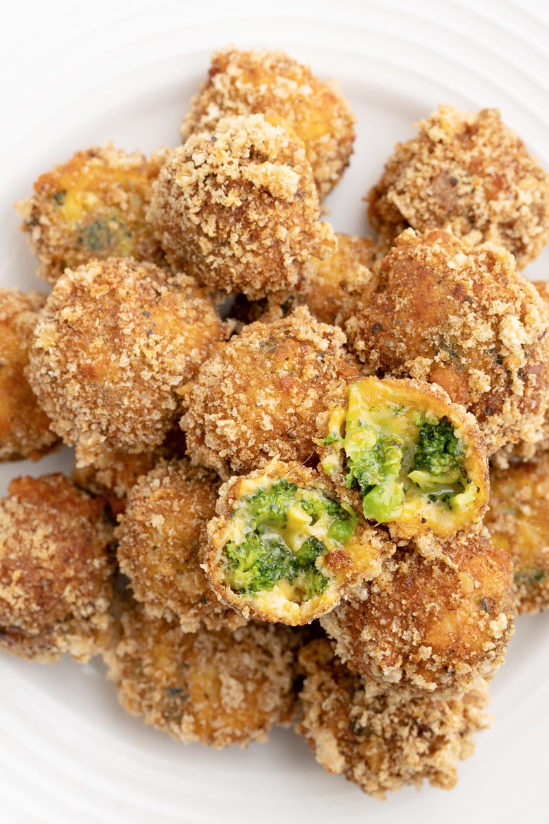 Close up of keto broccoli cheese balls piled on a white plate with one cheese ball cut open showing gooey cheese inside.