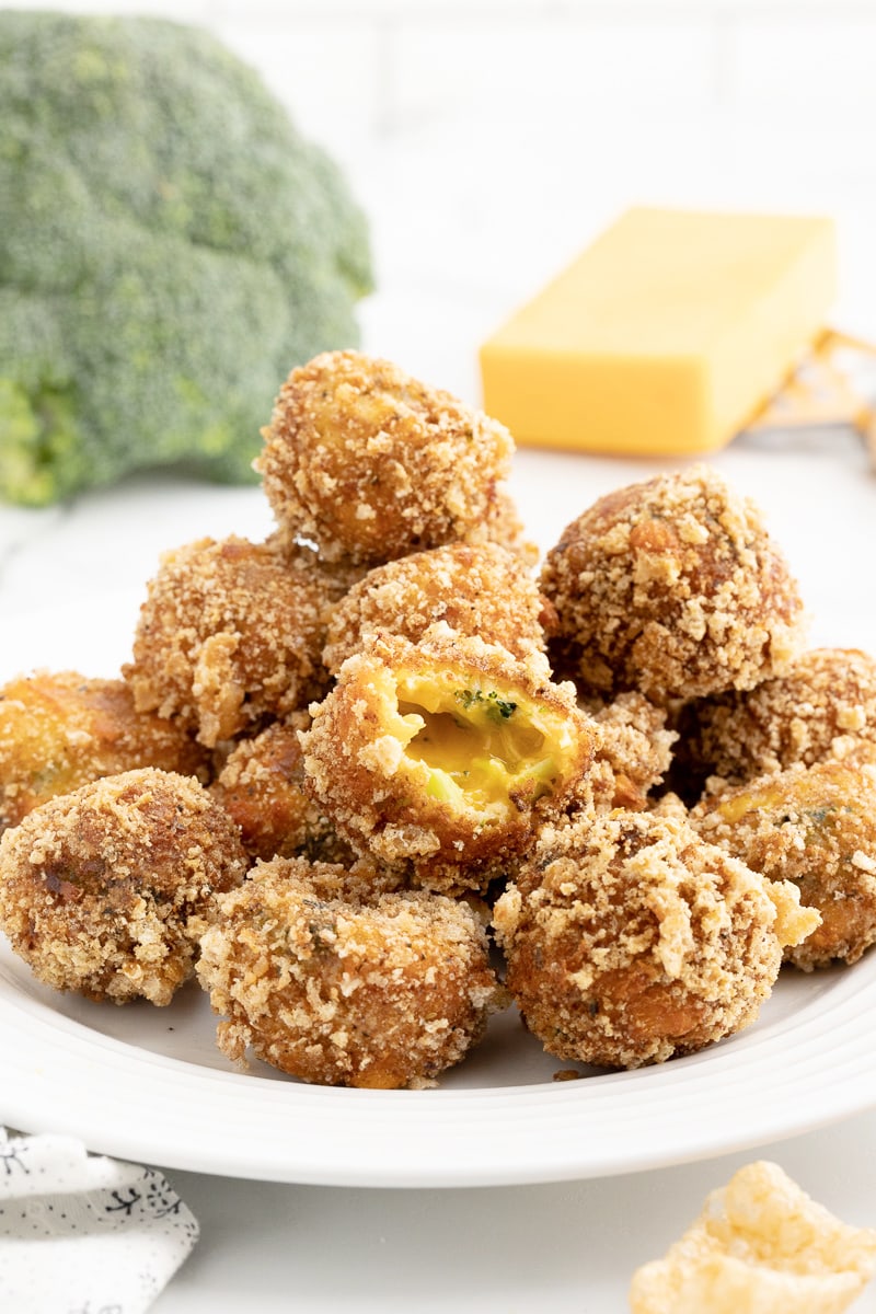 Keto broccoli cheeseballs piled up on a white plate with a head of broccoli and a block of cheese in the background.