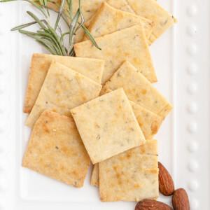 Keto garlic herb crackers on a white serving tray with a sprig of rosemary and some almonds.