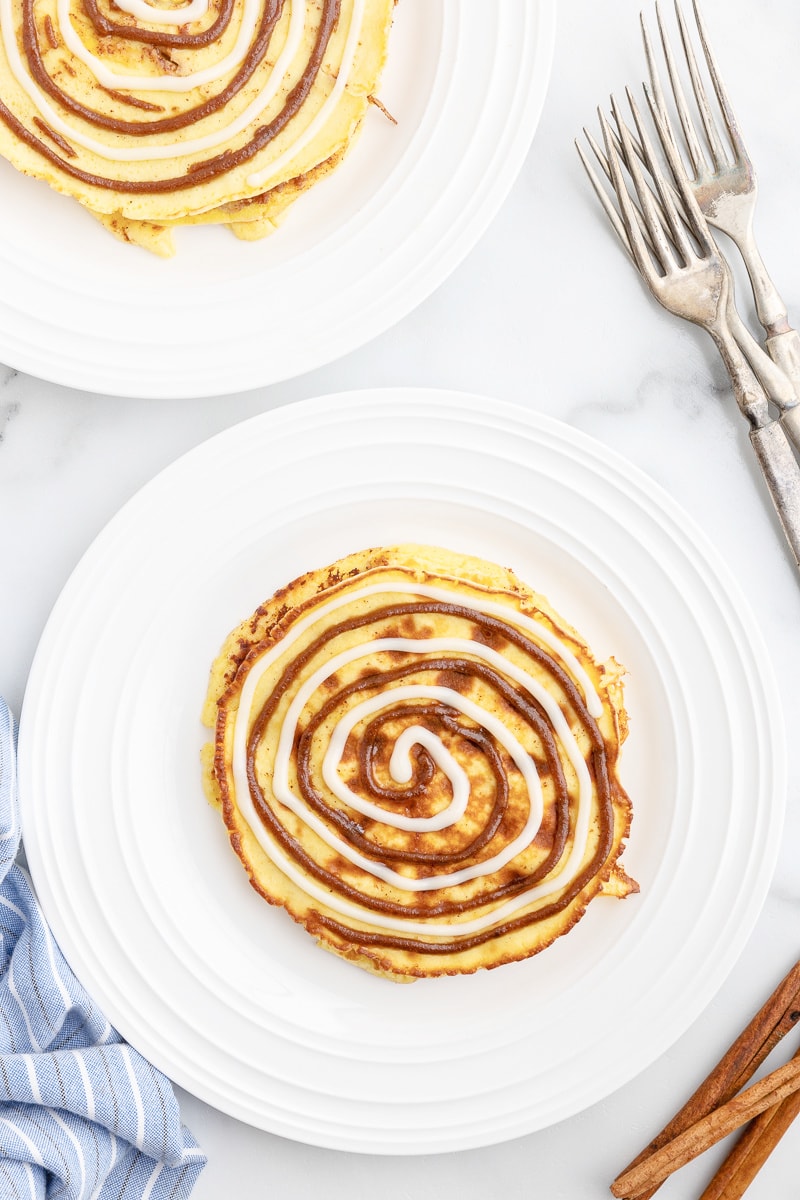 Overhead view of keto cinnamon roll pancakes on white plates with silver forks on the counter next to them.