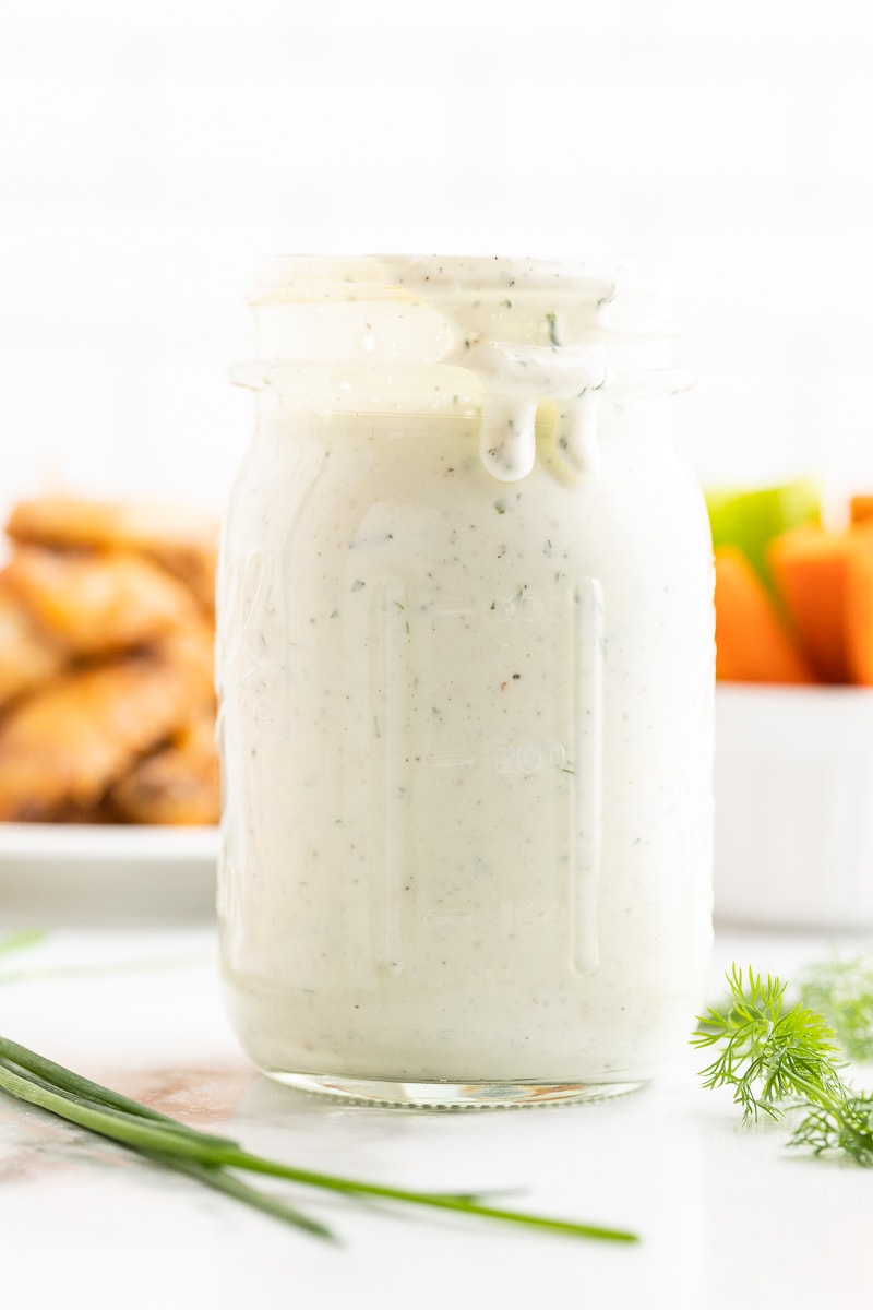 Homemade ranch dressing in a mason jar on a white counter with chicken wings and carrot and celery sticks in the background.