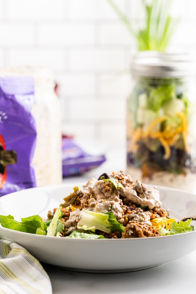 The taco salad from the mason jar in a white salad bowl with a bag of keto taco flavored chips and an other jar of salad in the background.