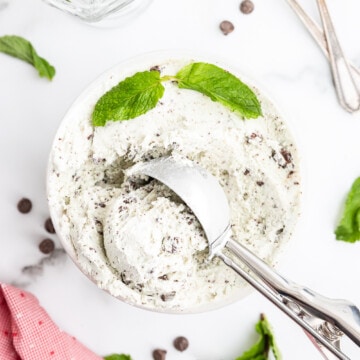 Keto mint chocolate chip ice cream in a tub with an ice cream scoop on a white marble counter.