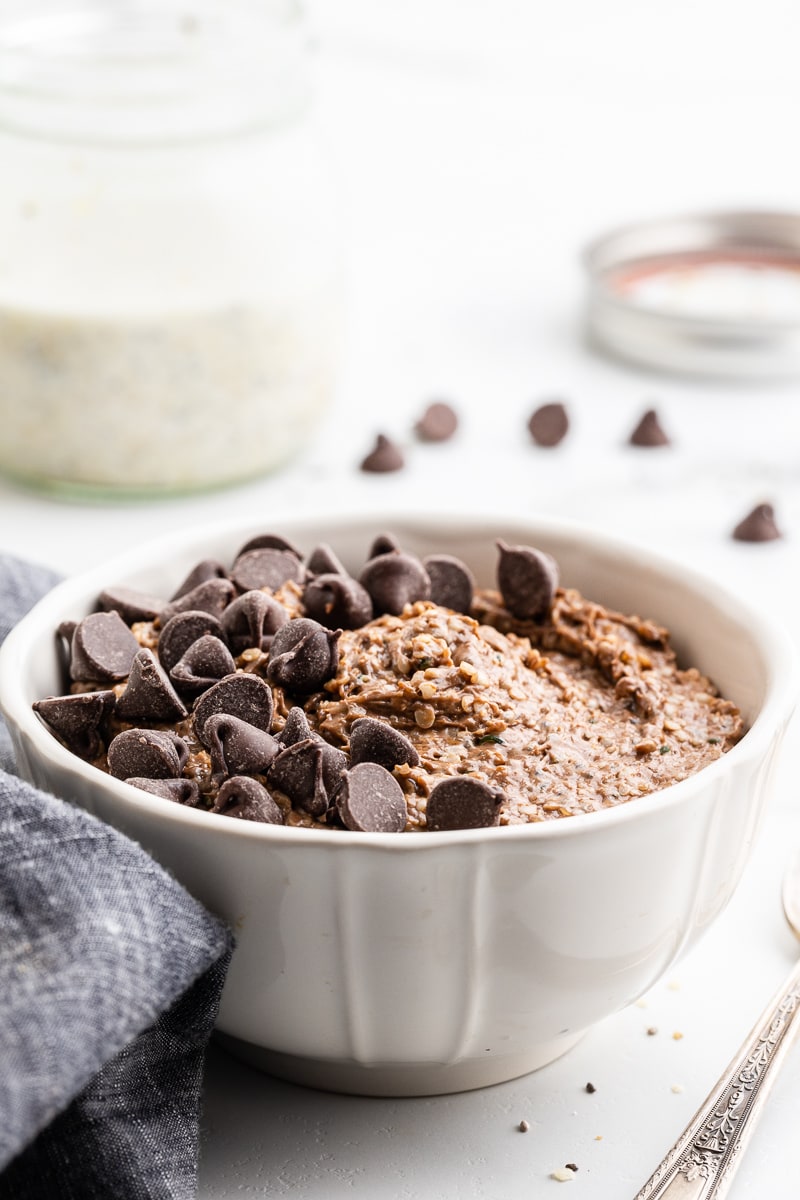 Side view photo of a white bowl on a gray surface, filled with keto double chocolate oatmeal. The oatmeal is topped with small chunks of dark chocolate and a silver spoon is placed to the right of the bowl. In the background, there is a small mason jar and a gray towel on the left side.
