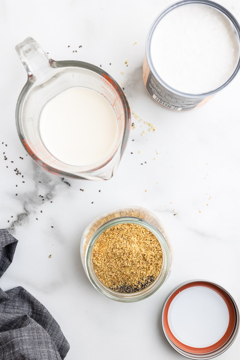Overhead photo of ingredients on a white marble counter for making keto oatmeal, including an open can of coconut milk, a glass measuring cup with heavy cream, and dry ingredients in a small mason jar. Chia seeds are scattered on the counter, and a gray towel and mason jar lid are in the corners.