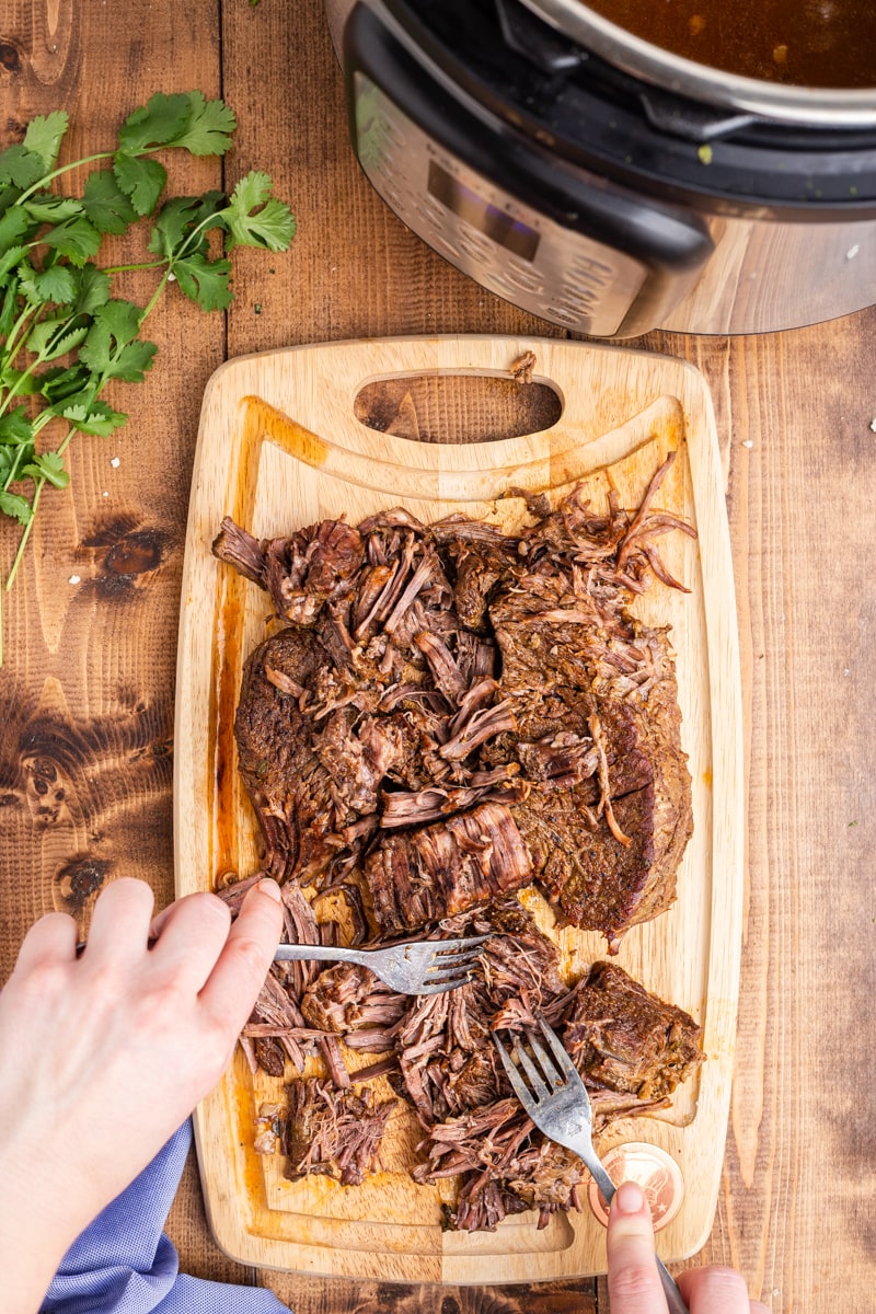 Shredding a cooked beef roast on a wooden cutting board with two forks.