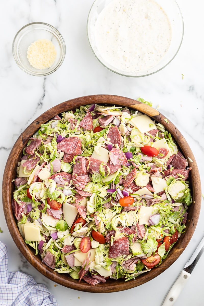 The shredded Brussels sprouts, salami, ham, provolone cheese, red onion, and tomatoes needed to make an Italian Grinder Salad a large wooden salad bowl on a white marble counter with a glass bowl container the dressing above it and to the right.