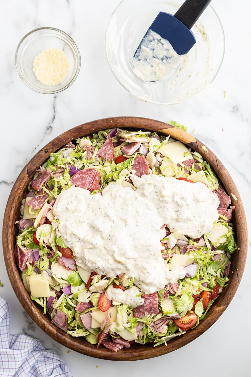 The shredded Brussels sprouts, salami, ham, provolone cheese, red onion, and tomatoes needed to make an Italian Grinder Salad with the homemade dressing poured on top in a large wooden salad bowl on a white marble counter