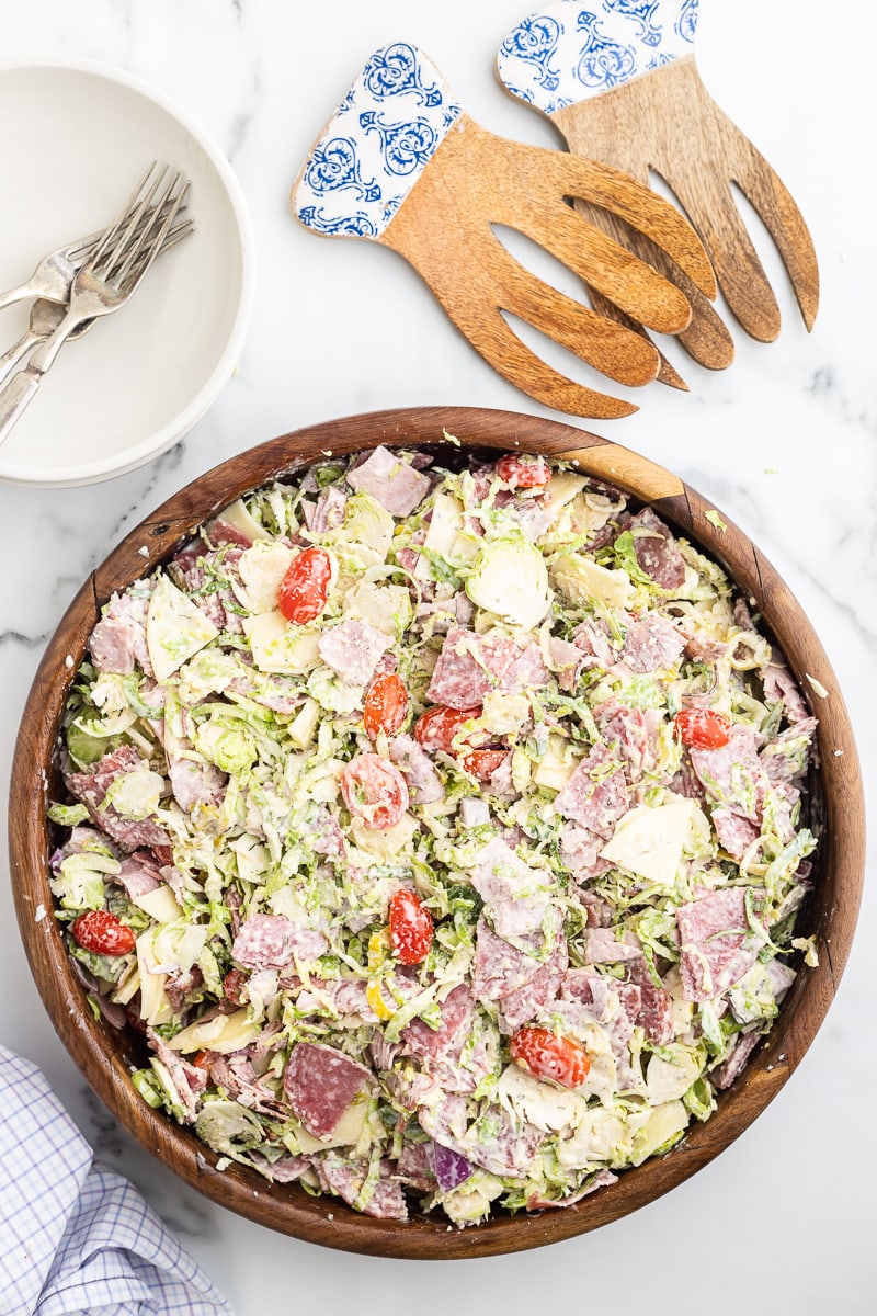 The shredded Brussels sprouts, salami, ham, provolone cheese, red onion, and tomatoes needed to make an Italian Grinder Salad combined with the homemade dressing in a large wooden salad bowl on a white marble counter