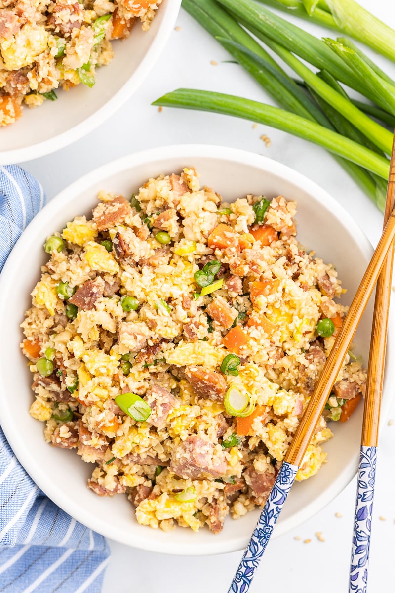 Overhead view of ham fried cauliflower rice in a white bowl with wooden chopsticks.