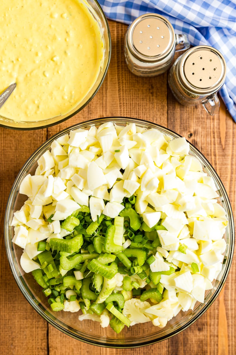 Chopped vegetables and egg whites for deviled egg cauliflower salad in a large glass bowl at the center of the photo. A medium glass bowl with mayo, mustard, pickle relish, and mashed egg yolks for the sauce is to the upper left. In the upper right, a salt and pepper shaker and a blue and white checkered kitchen towel are visible on a wooden table.