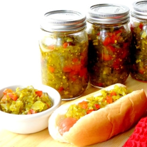 Mason jars of spicy pickle relish and a white bowl full of relish with a hot dog topped with the relish on a wooden cutting board.