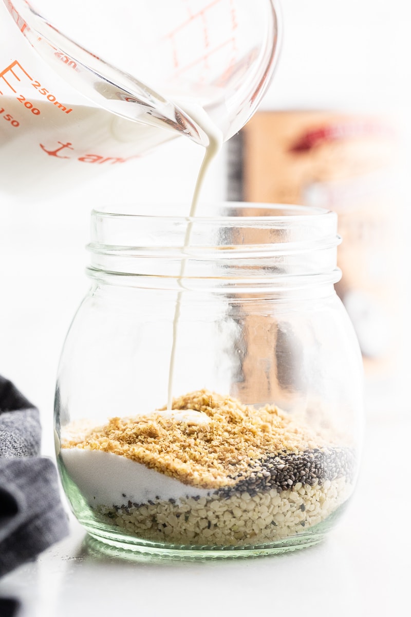 Glass measuring cup pouring heavy cream and coconut milk into small mason jar with dry ingredients for keto oatmeal. Layered view of ingredients.