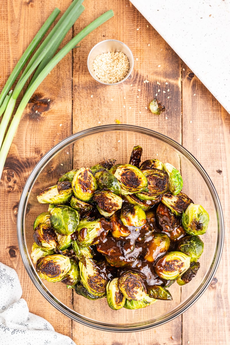 Pouring Kung Pao sauce over Brussels sprouts in a glass bowl.