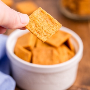 A white male presenting hand holding a keto smoked cheddar cheez-it cracker over a white ramekin full of them.