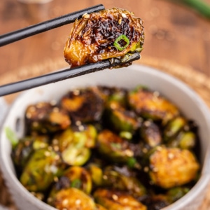 Black chopsticks holding a Kung Pao Brussels Sprout over a white bowl full of them.