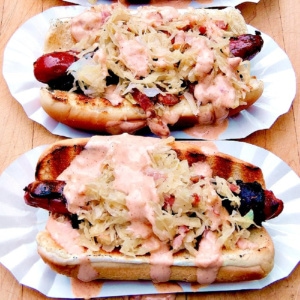 Close up of 2 Reuben Hot Dogs on white paper hot dog holders.