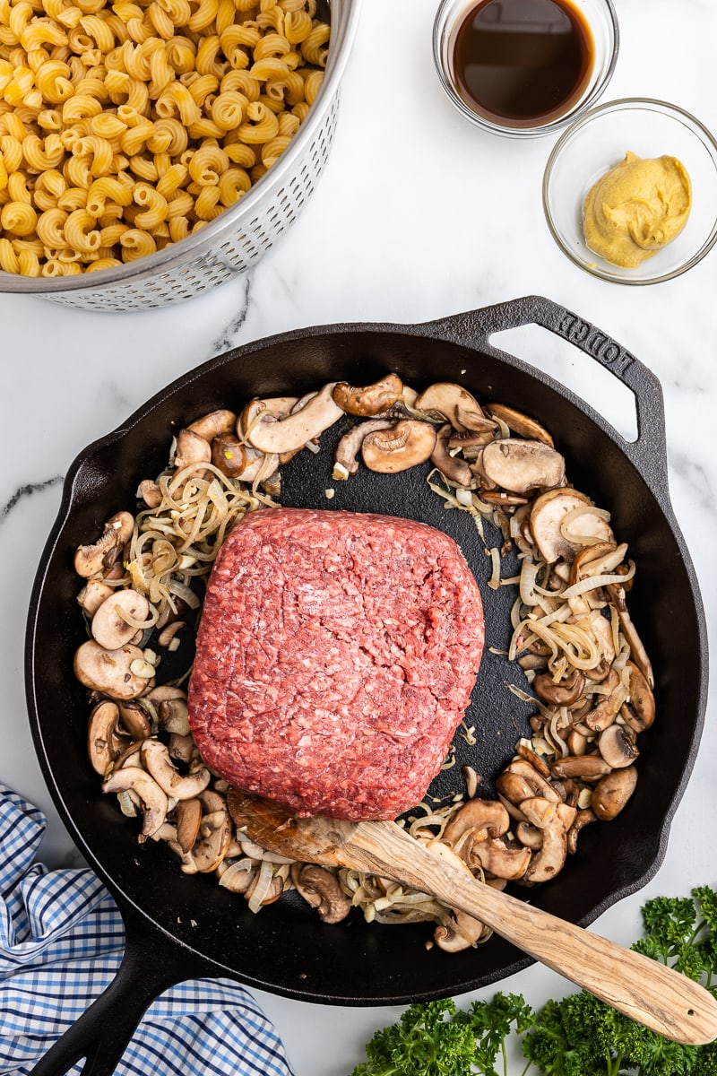 Cooked onions and mushrooms pushed to the side of a cast iron skillet and 1 pound of ground beef in the center.