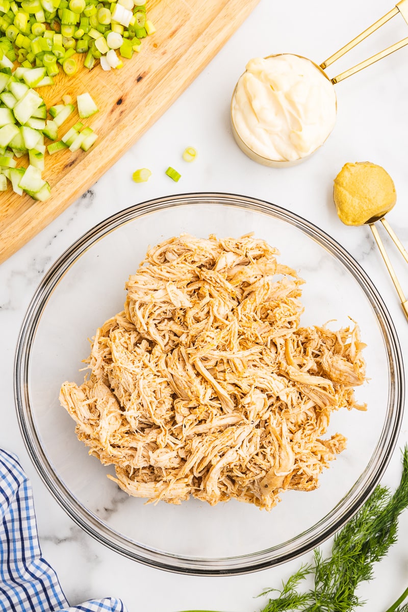 Overhead view of shredded cooked chicken in a glass bowl on a white marble counter with fresh dill, chopped cucumber and green onion, a measuring cup with mayo and measuring spoon with mustard beside the bowl.