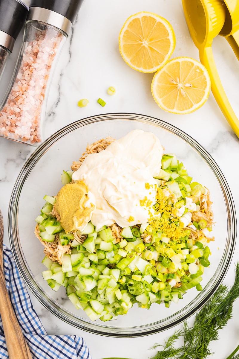 Overhead view of shredded cooked chicken, chopped cucumber and green onions, mayo, mustard, and lemon zest in a glass bowl on a white marble counter with fresh dill, a cut lemon and citrus juicer, and salt and pepper shakers next to it.