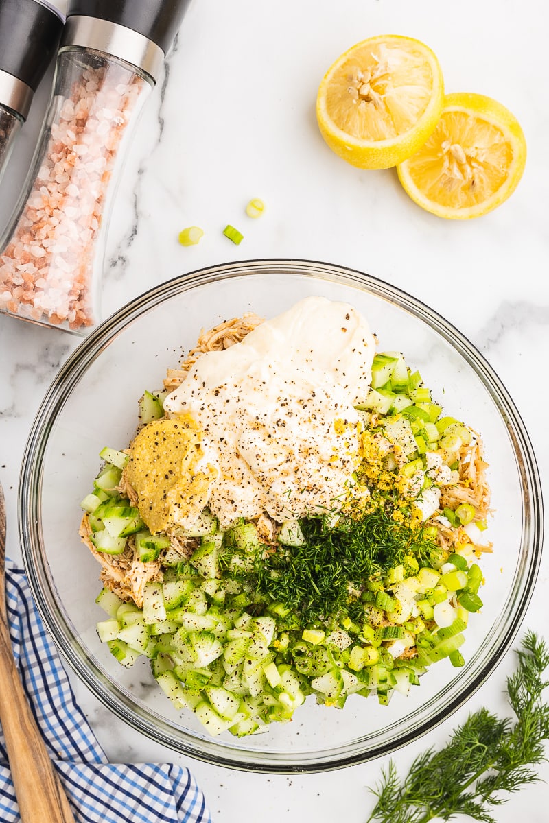 Overhead view of shredded cooked chicken, chopped cucumber and green onions, mayo, mustard, lemon zest, fresh dill, and salt and pepepr in a glass bowl on a white marble counter.