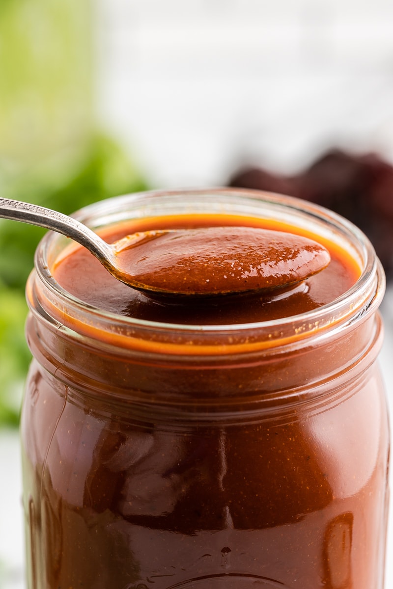Homemade red enchilada sauce in a glass jar with a silver spoon holding a spoonful of sauce above it for a 60 Drool-worthy Keto and Low Carb Mexican Recipes collection.