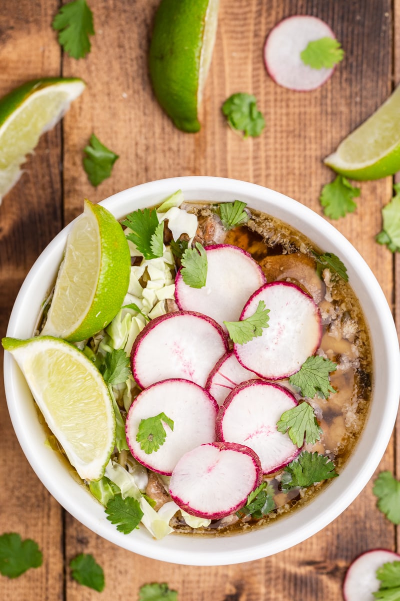 Overhead view of a white bowl filled with keto pork pozole verde on a wooden table. The soup is topped with sliced cabbage, sliced radish, fresh cilantro, and a wedge of lime.