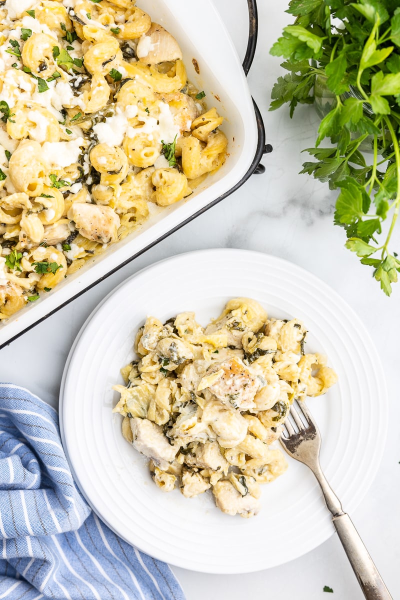 Keto Spinach Artichoke Chicken Casserole served on a white plate and placed on a marble countertop. The casserole is baked to perfection with a golden brown bubbly cheese topping and has chunks of chicken, low-carb pasta, and spinach artichoke dip visible.