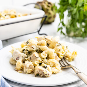 Keto Spinach Artichoke Chicken Casserole served on a white plate and placed on a marble countertop. The casserole is baked to perfection with a golden brown bubbly cheese topping and has chunks of chicken, low-carb pasta, and spinach artichoke dip visible.
