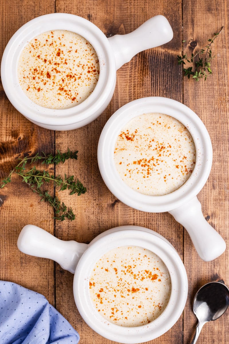 A wooden table with three white crocks filled with low carb keto oyster stew, each bowl has a handle and is topped with a sprinkle of paprika.