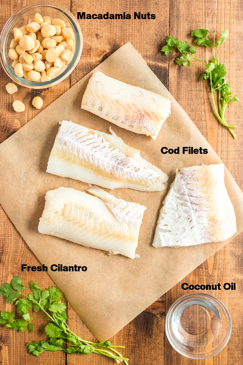 Overhead photo of ingredients needed to make Oven Baked Coconut Macadamia Fish Fillets - macadamia nuts, cod fillets, coconut oil, and fresh cilantro arranged on a wooden table.