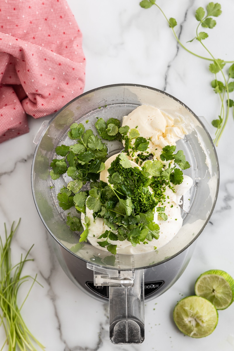 Overhead view of all of the ingredients needed to make Cilantro Lime Crema in the bowl of a food processor. 