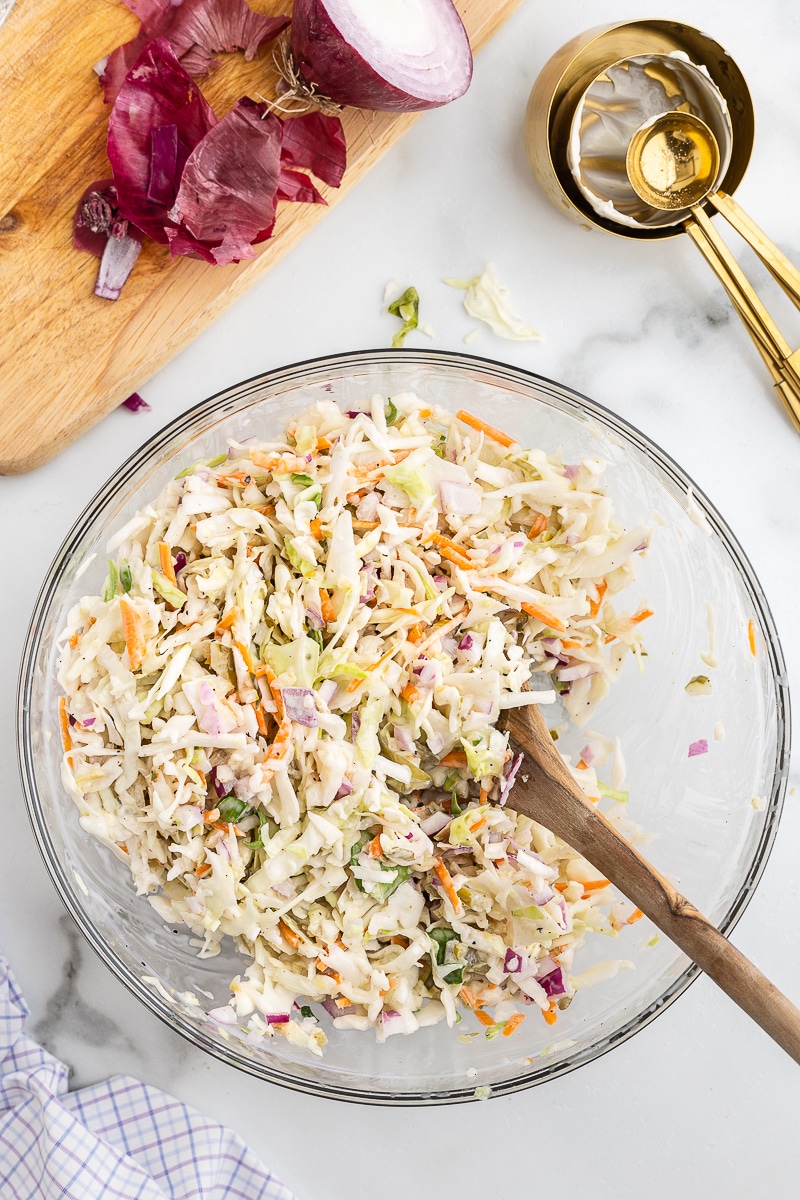 Overhead view of dill pickle coleslaw in a large glass mixing bowl with a wooden spoon.