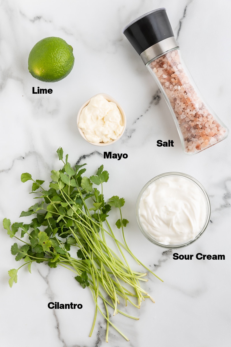Overhead view of the ingredients needed to make Cilantro Lime Crema on a white marble counter.