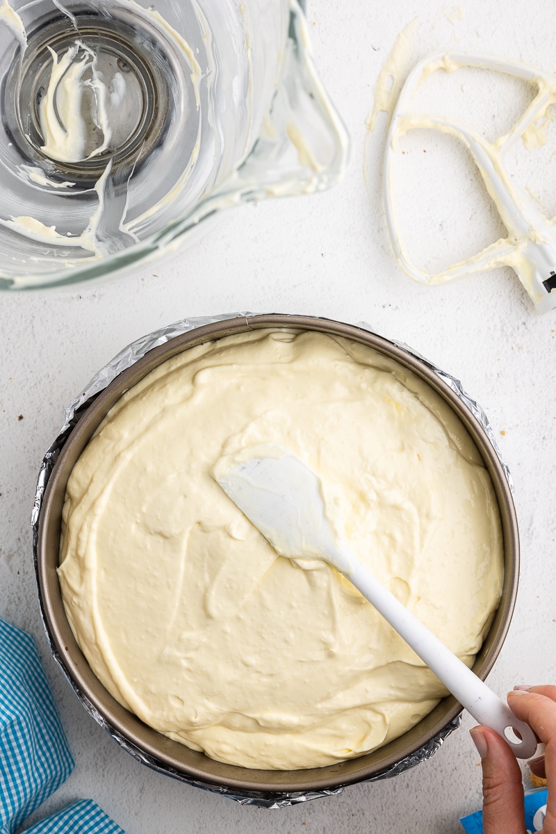 Overhead view of a white hand smoothing the cheesecake mixture into a springform pan on a white marble counter.